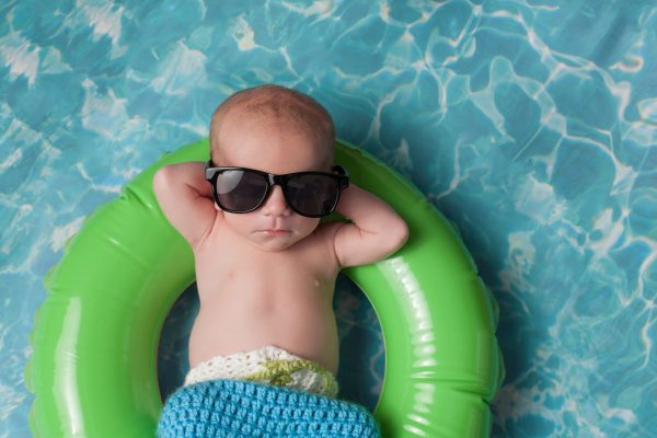 Four week old newborn baby boy sleeping on a tiny inflatable swim ring. He is wearing crocheted board shorts and black sunglasses.