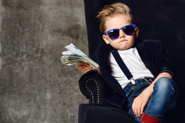 Happy and shoutting rich kid boy millionaire sits with a bundle of money dollars cash in big luxury armchair while bills falling